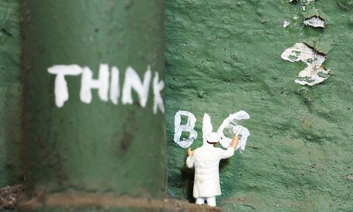 Photograph of a toy figurine dressed in white painting the words "think big" in white on a green wall